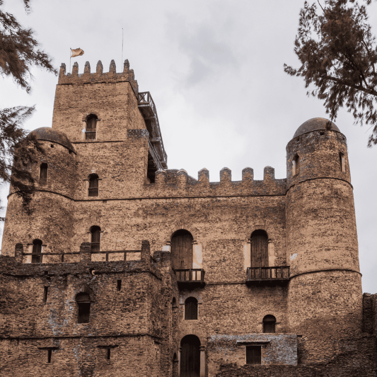 The stone castle of Fasilides at Gondar seen from below through pine trees, crenellated walls and domed corner towers against a pale sky