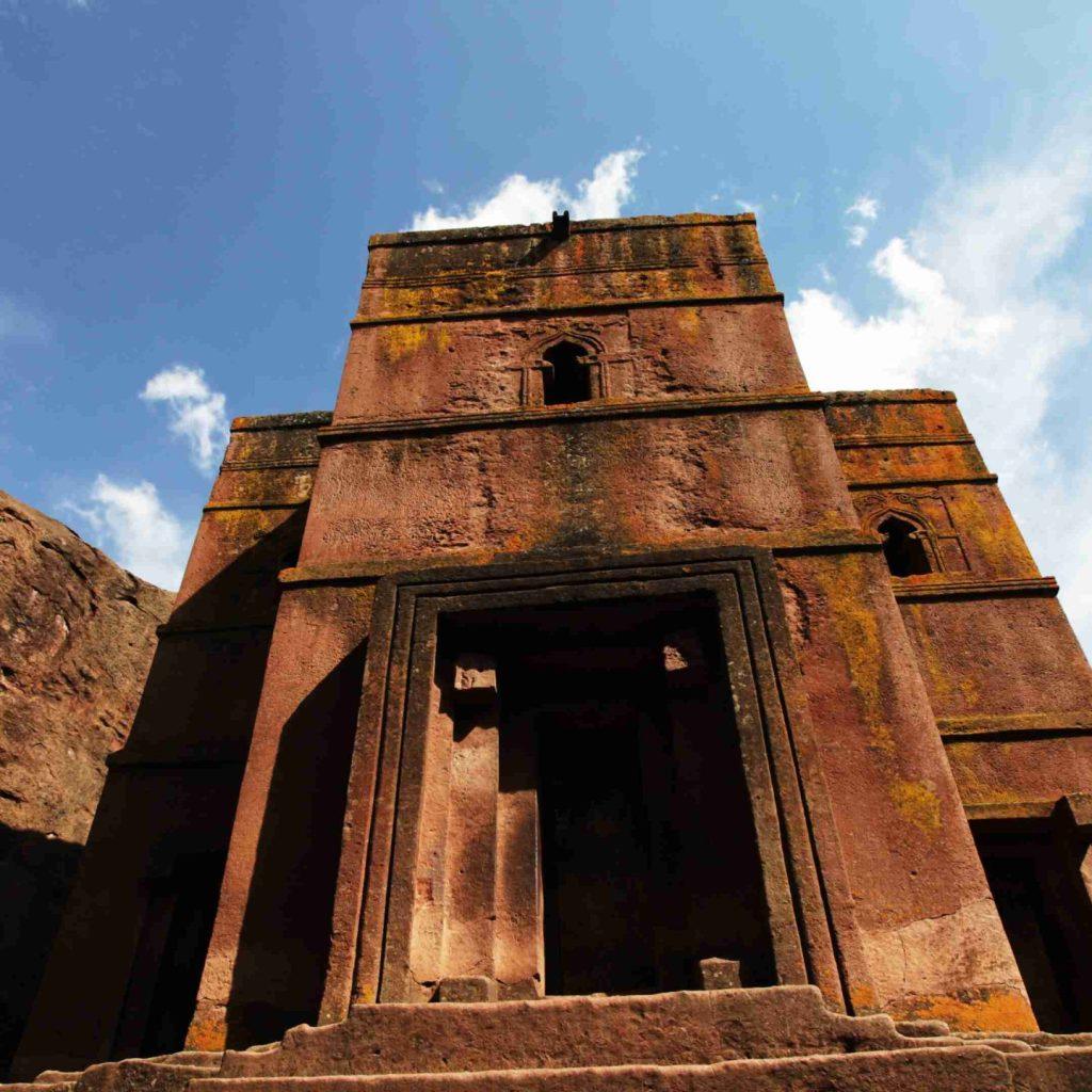 A rock-hewn church at Lalibela cut down into the red volcanic rock, its tiered facade and carved doorway seen from below against a blue sky