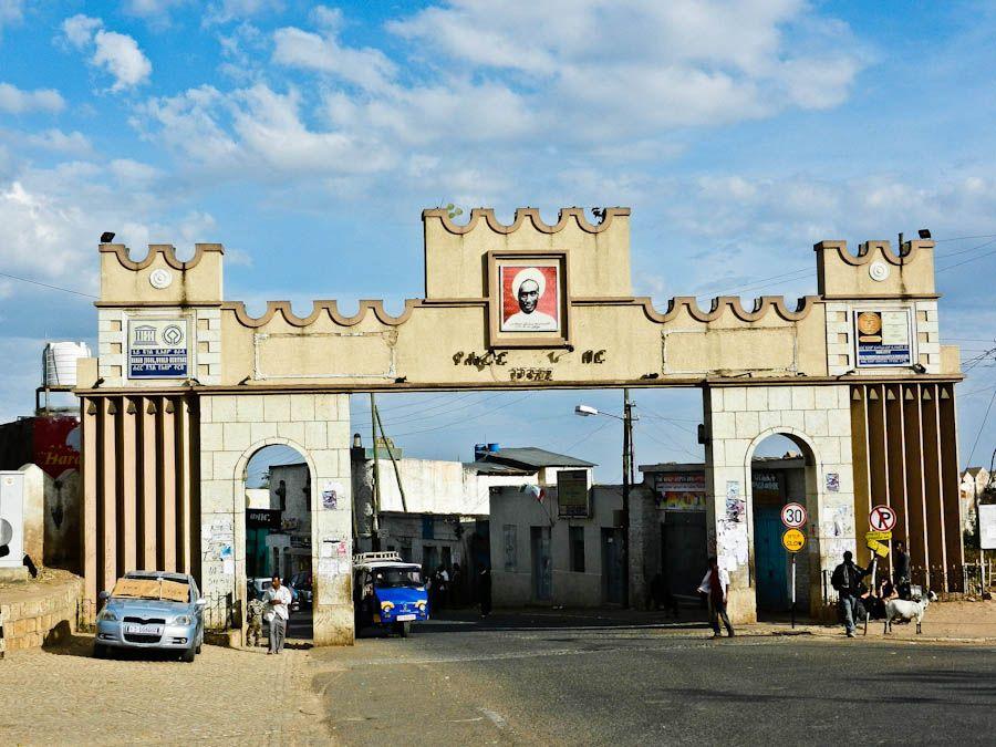 The crenellated Harar Gate with twin arches and a portrait plaque above, traffic and a blue three-wheeler passing beneath and goats at the roadside