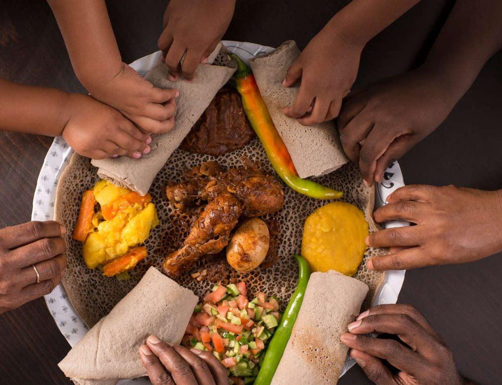 Six pairs of hands reaching in from all sides of a shared injera platter to tear pieces of bread, with doro wat chicken and a hard-boiled egg in the centre alongside potato, split pea, salad and green chillies