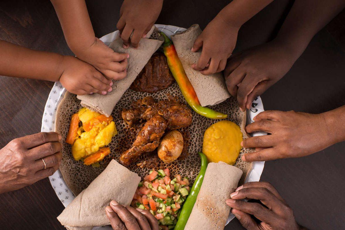 Six pairs of hands reaching in from all sides of a shared injera platter to tear pieces of bread, with doro wat chicken and a hard-boiled egg in the centre alongside potato, split pea, salad and green chillies