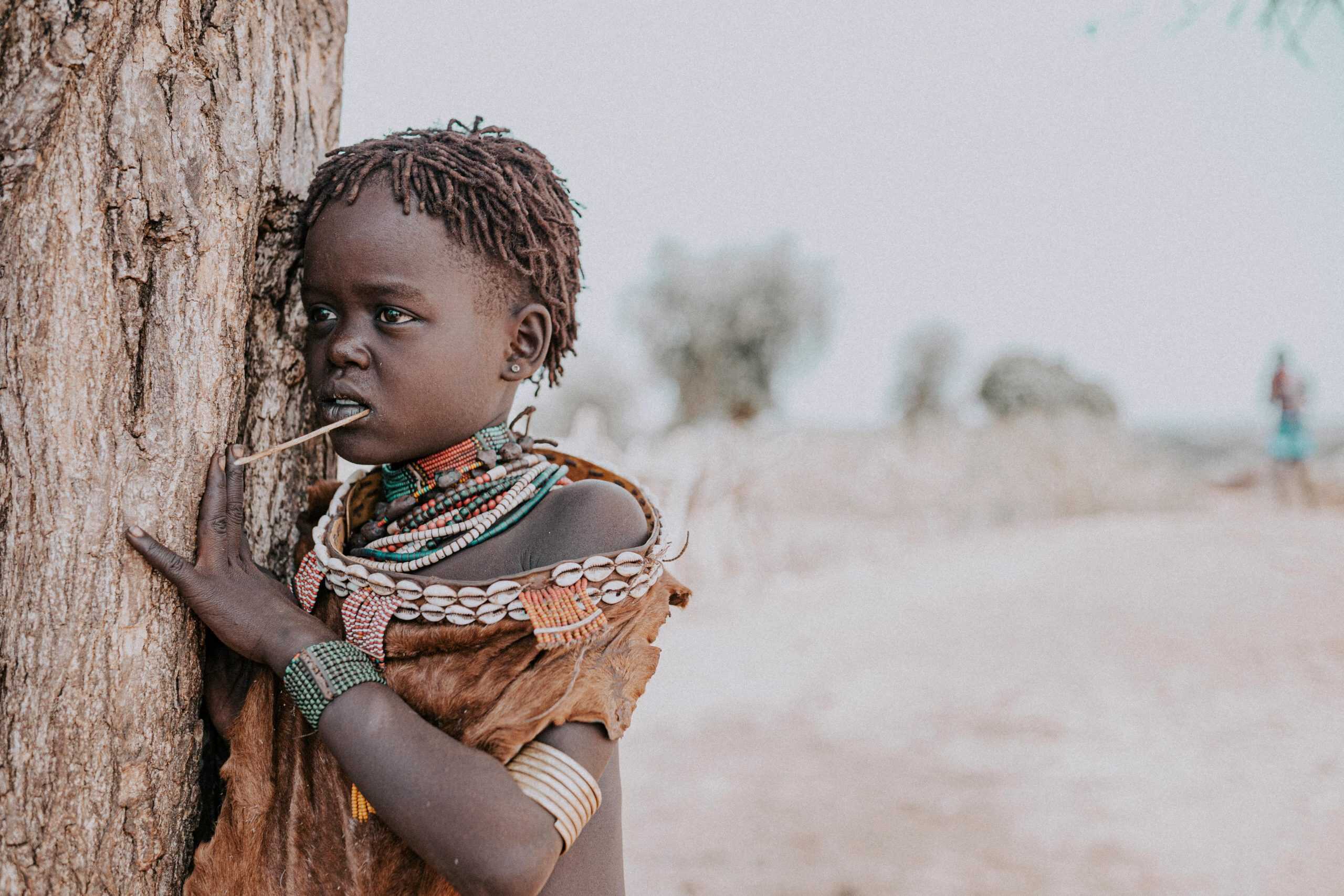 A Hamar child in beaded necklaces and cowrie shells standing beside a tree in the Omo Valley