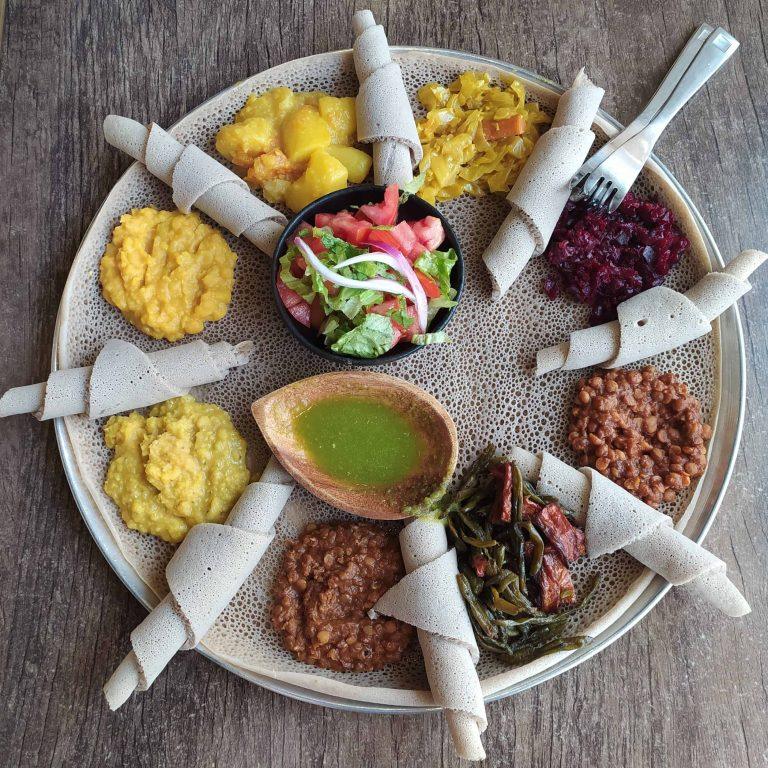 An Ethiopian fasting platter seen from above, a large round injera covered with mounds of yellow split pea, red lentil stew, collard greens, potato and carrot, beetroot and fresh salad, with rolled pieces of injera arranged around the edge