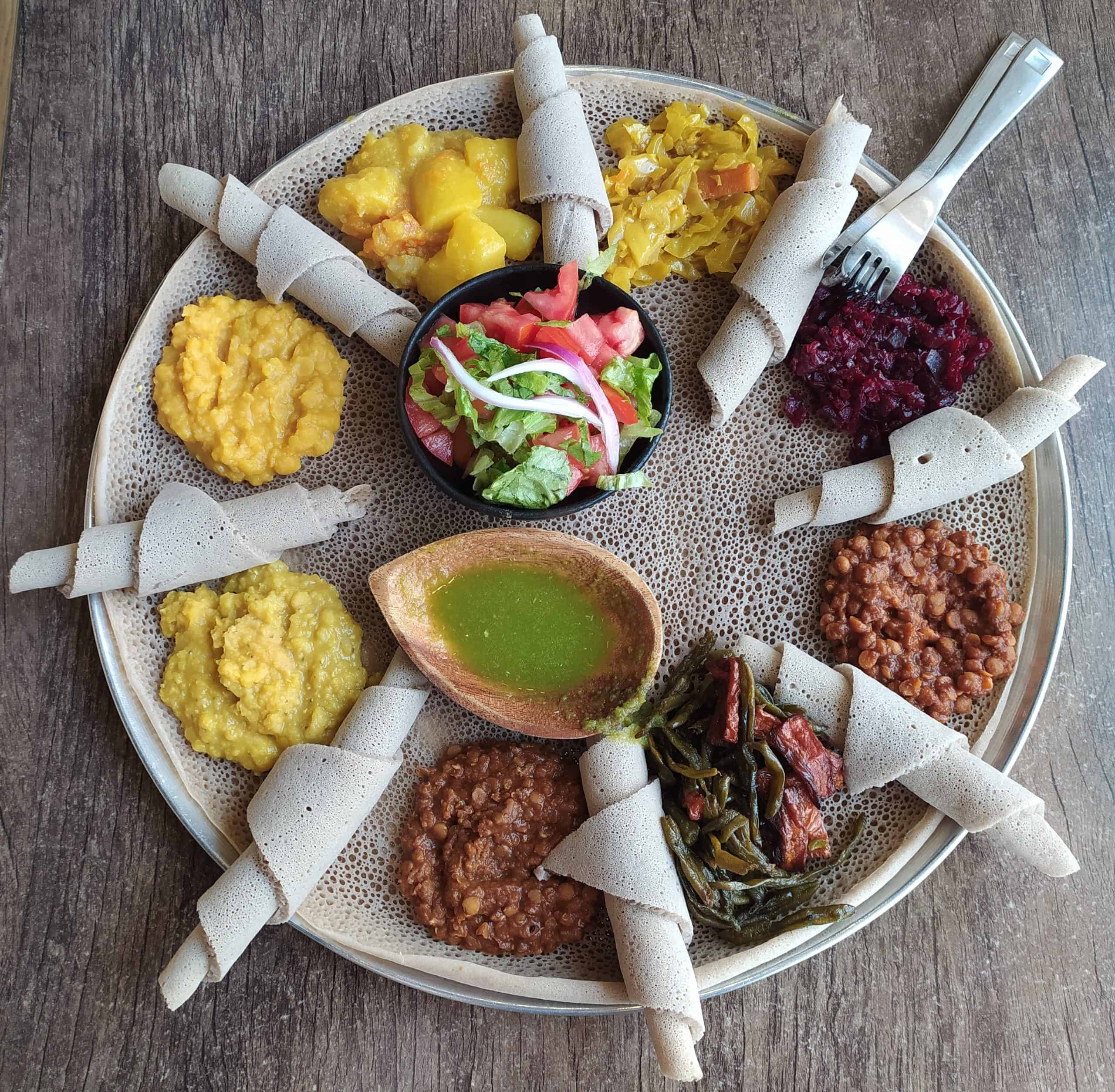 An Ethiopian fasting platter seen from above, a large round injera covered with mounds of yellow split pea, red lentil stew, collard greens, potato and carrot, beetroot and fresh salad, with rolled pieces of injera arranged around the edge