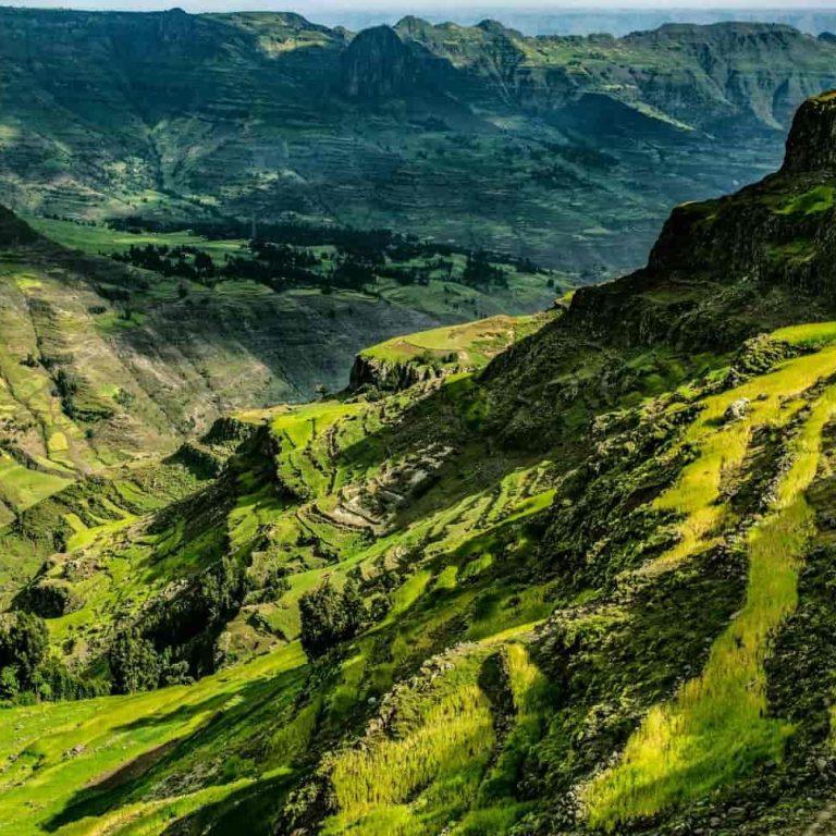 Green terraced farmland on steep Ethiopian highland slopes, with layered basalt cliffs and deep valleys receding into haze and a few thatched dwellings at the foot of the slope