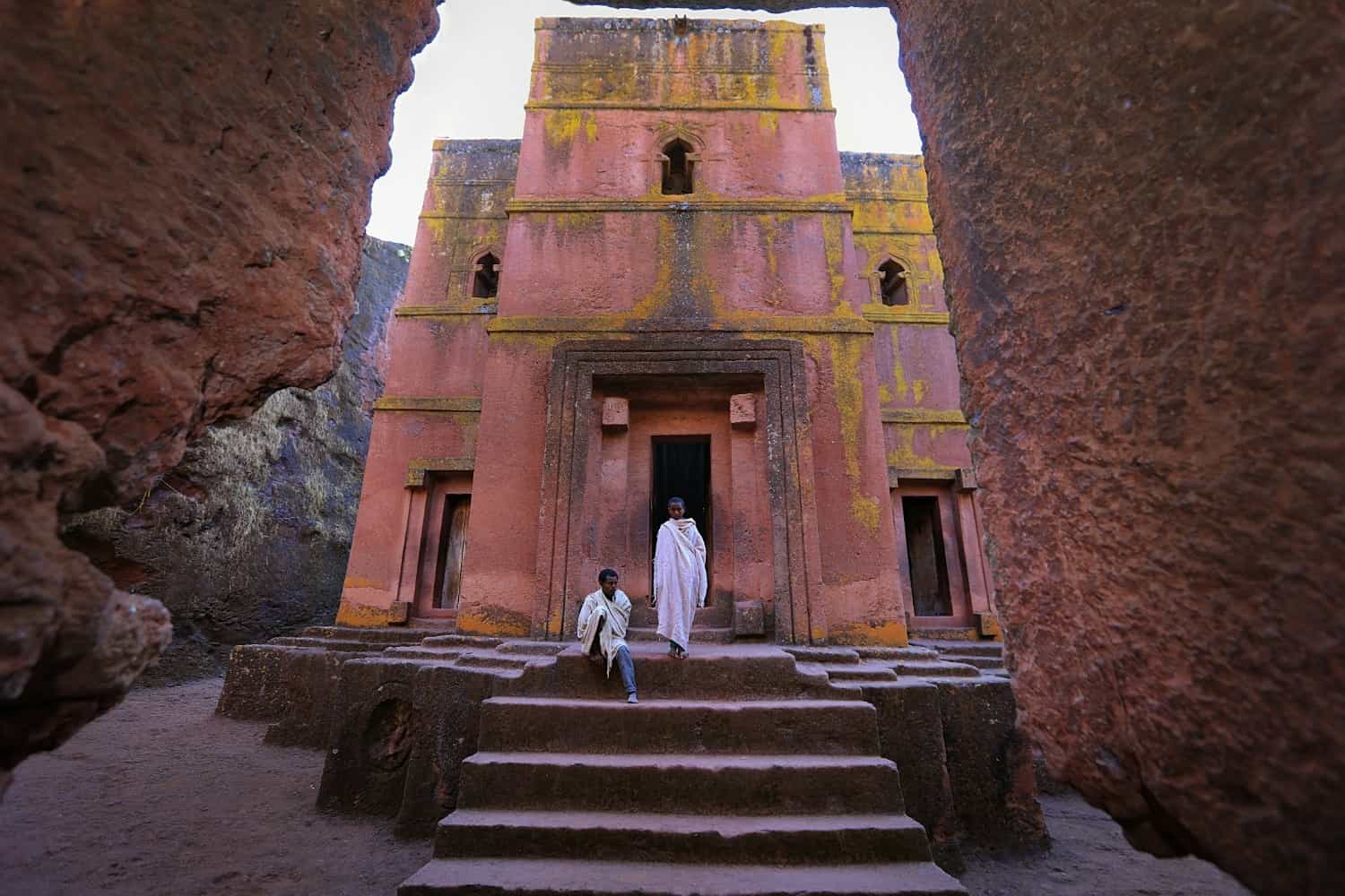 Two people in white robes walking down the carved rock steps in front of Bete Giyorgis at Lalibela, the cross-shaped church cut down into pink-red volcanic rock streaked with yellow lichen, seen from inside the surrounding trench