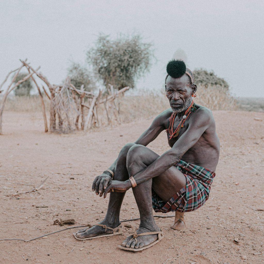 An older man sitting on bare red earth in a checked cloth wrap and beaded necklaces, his hair dressed in a clay bun with a feather, a branch shelter behind him