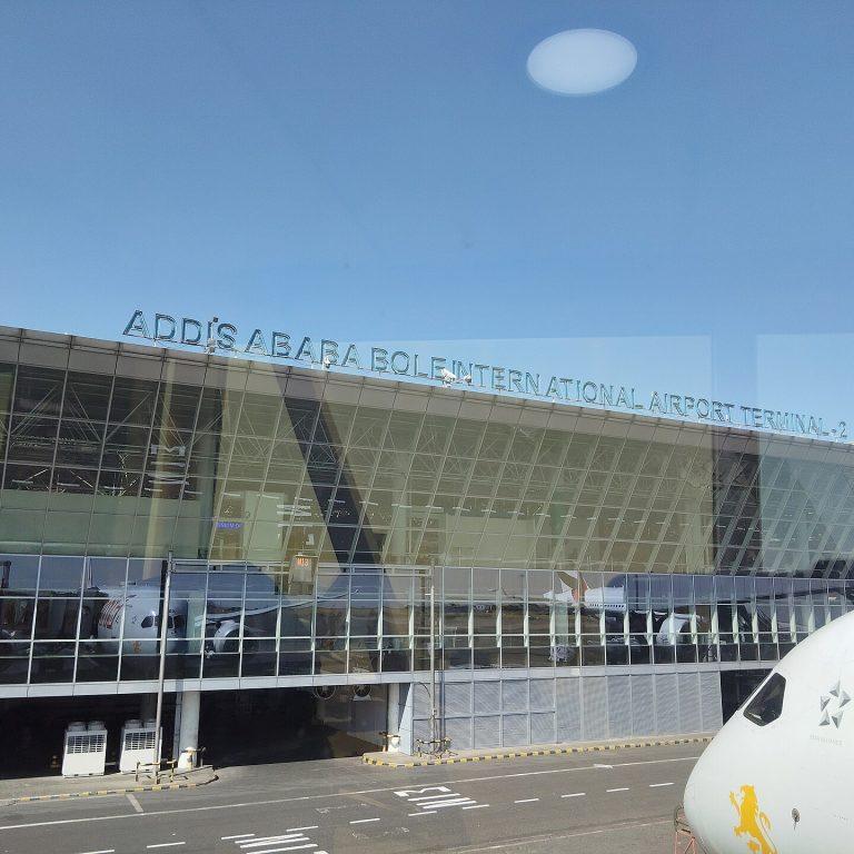 The glass frontage of Addis Ababa Bole International Airport Terminal 2, with the terminal name across the roofline and the nose of an Ethiopian Airlines aircraft on the apron alongside