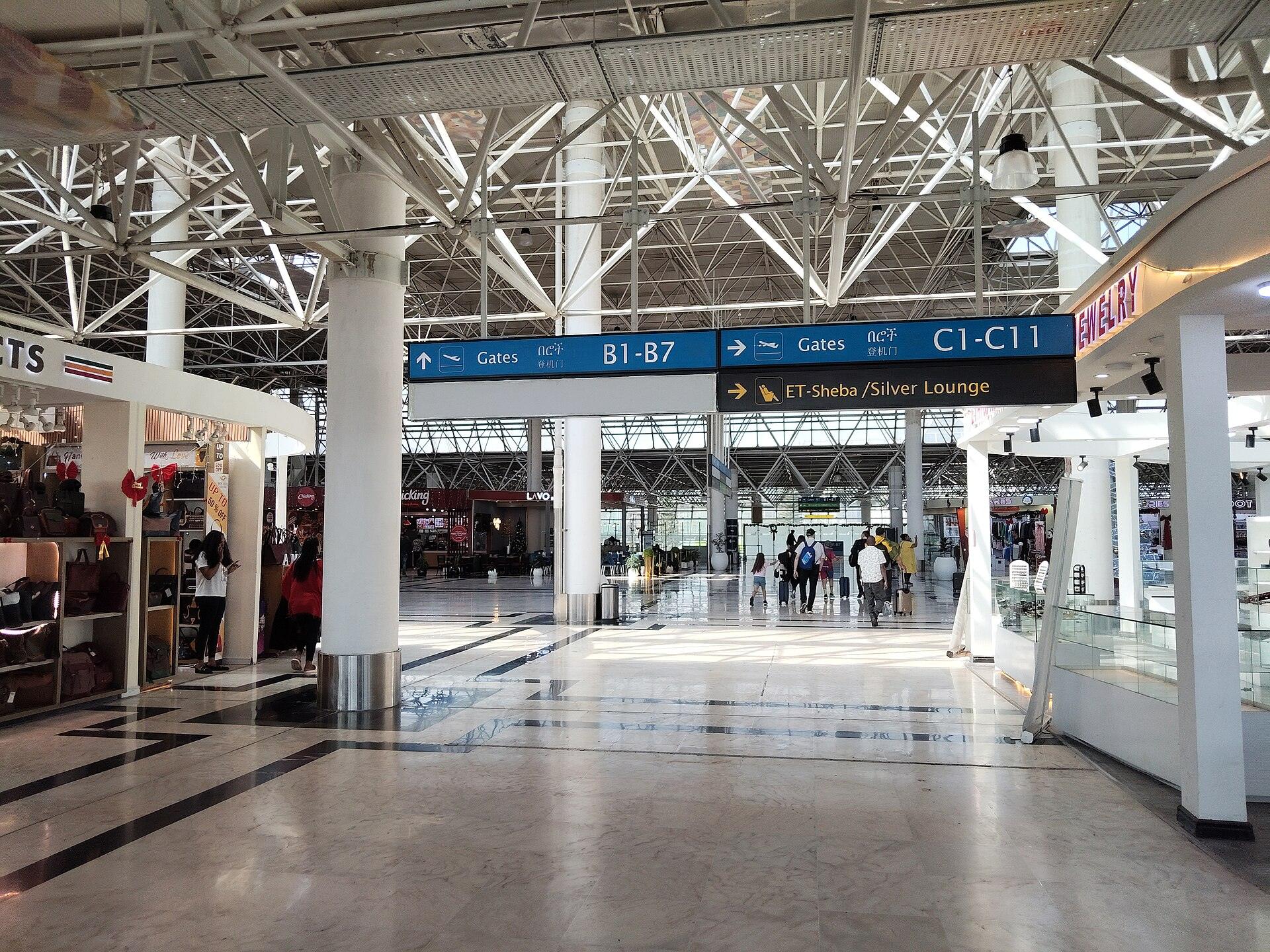 Overhead direction signs inside Bole airport pointing left to Gates B1 to B7 and right to Gates C1 to C11, with passengers walking through the concourse with wheeled luggage