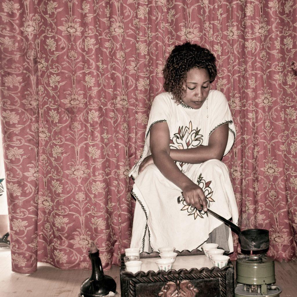 A woman in a white embroidered habesha dress roasting coffee beans in a long-handled pan over a burner, with a black clay jebena and a tray of small white cups in front of her