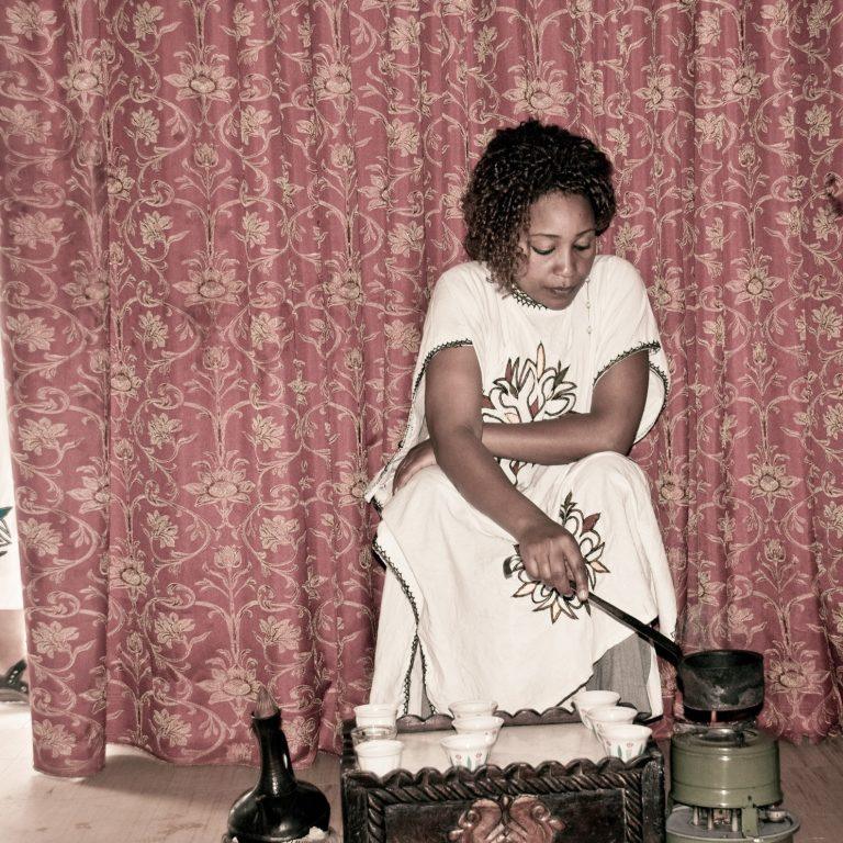 A woman in a white embroidered habesha dress roasting coffee beans in a long-handled pan over a burner, with a black clay jebena and a carved wooden table of small white cups in front of her