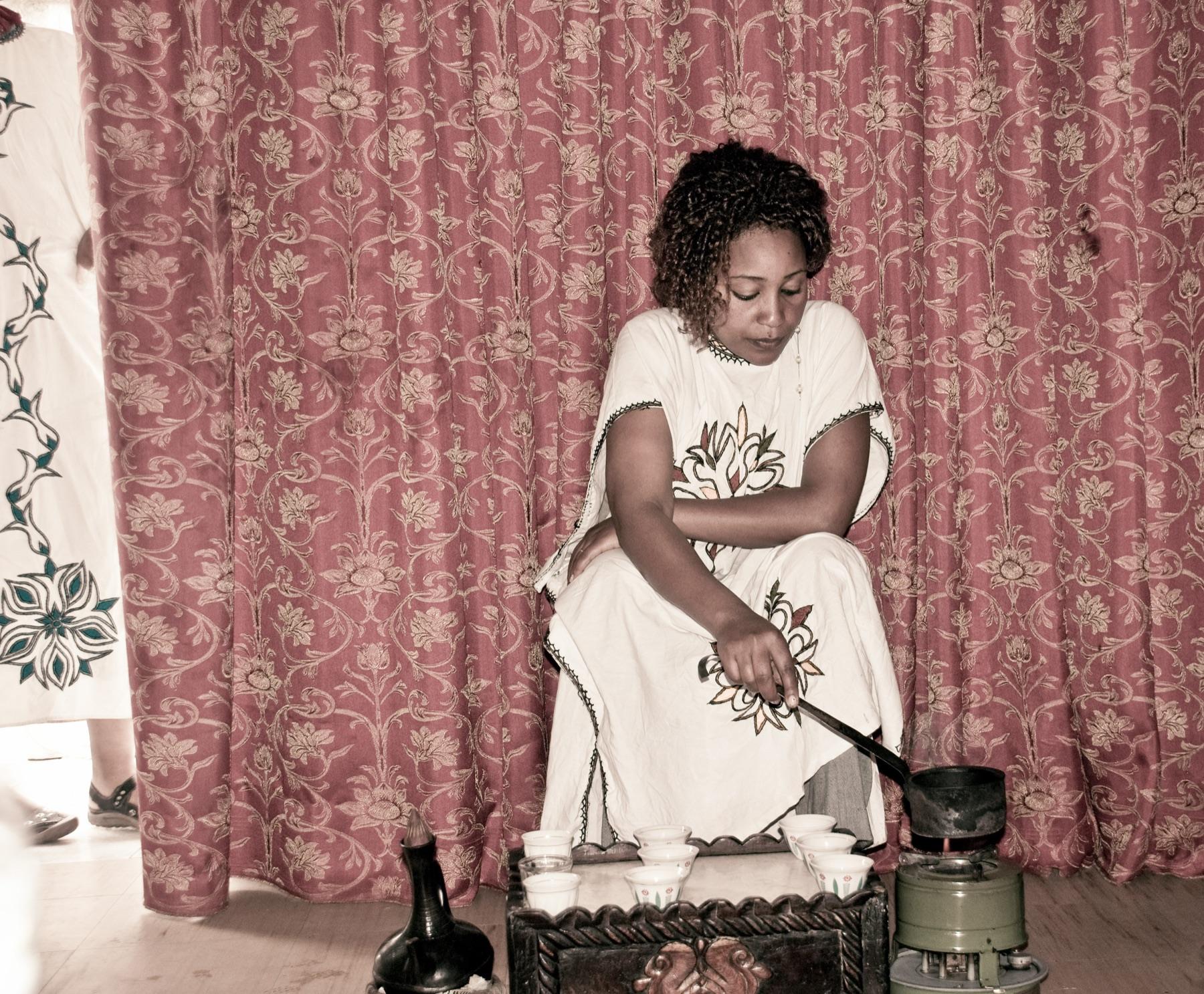 A woman in a white embroidered habesha dress roasting coffee beans in a long-handled pan over a burner, with a black clay jebena and a tray of small white cups in front of her