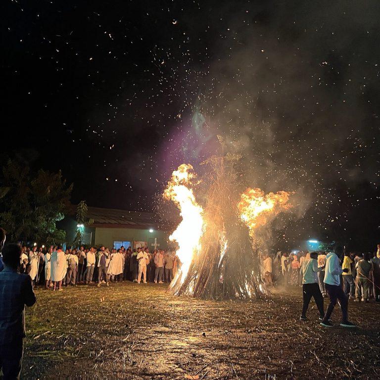 A tall conical demera bonfire burning at night with sparks rising into the dark, a crowd standing around it and a church building lit behind
