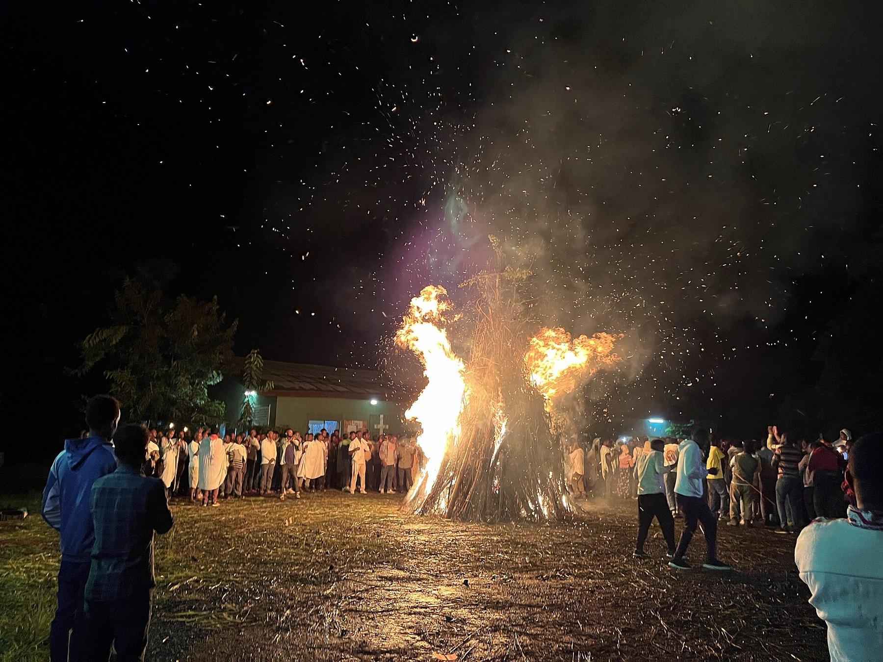 A tall conical demera bonfire burning at night with sparks rising into the dark, a crowd standing around it and a church building lit behind