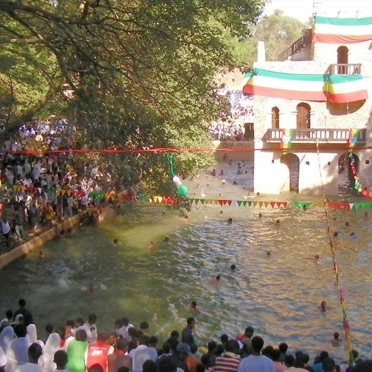 Crowds in white gathered around the flooded stone pool at Fasilides Bath during Timkat, with the pavilion draped in Ethiopian flag colours and people swimming in the water