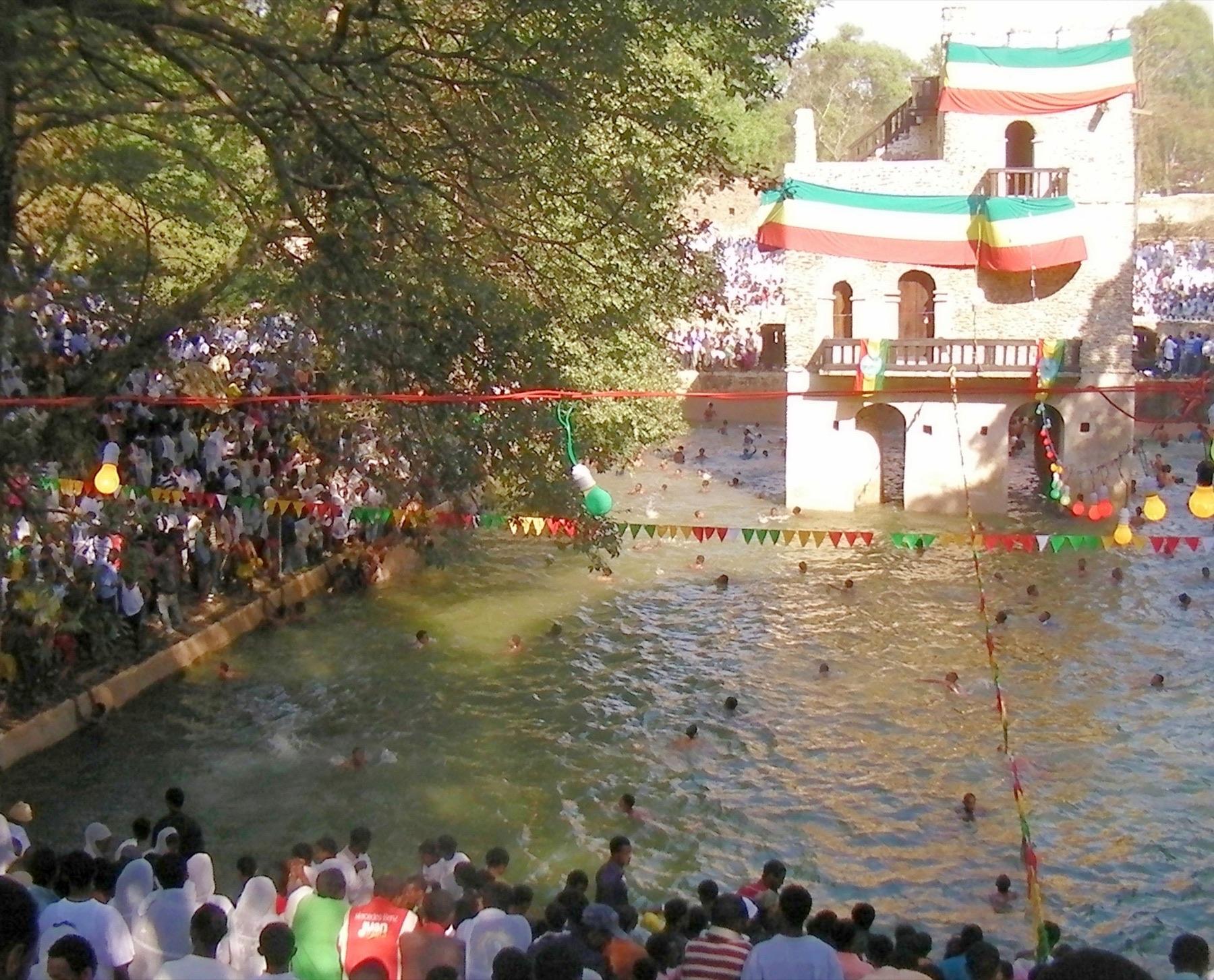 Crowds in white gathered around the flooded stone pool at Fasilides Bath during Timkat, with the pavilion draped in Ethiopian flag colours and people swimming in the water