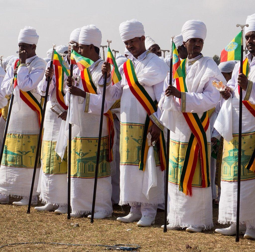 A long row of Ethiopian Orthodox priests in white robes with yellow and green embroidered borders and white turbans, each holding a prayer staff and a small Ethiopian flag