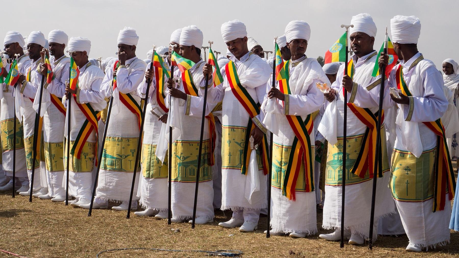 A long row of Ethiopian Orthodox priests in white robes with yellow and green embroidered borders and white turbans, each holding a prayer staff and a small Ethiopian flag