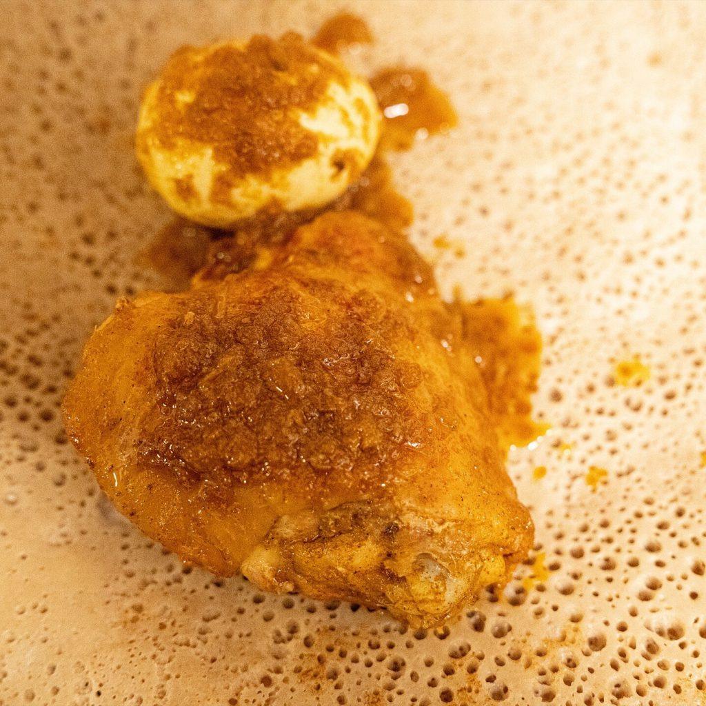 A piece of chicken in deep red berbere sauce with a hard-boiled egg beside it, served on injera whose fermentation holes are clearly visible