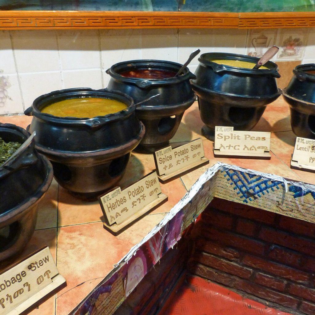 A row of black clay pots on stands holding split pea, lentil, potato and greens stews, each with a small wooden label in English and Amharic