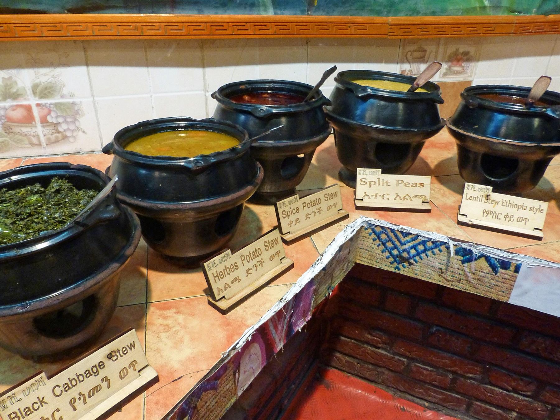 A row of black clay pots on stands holding split pea, lentil, potato and greens stews, each with a small wooden label in English and Amharic