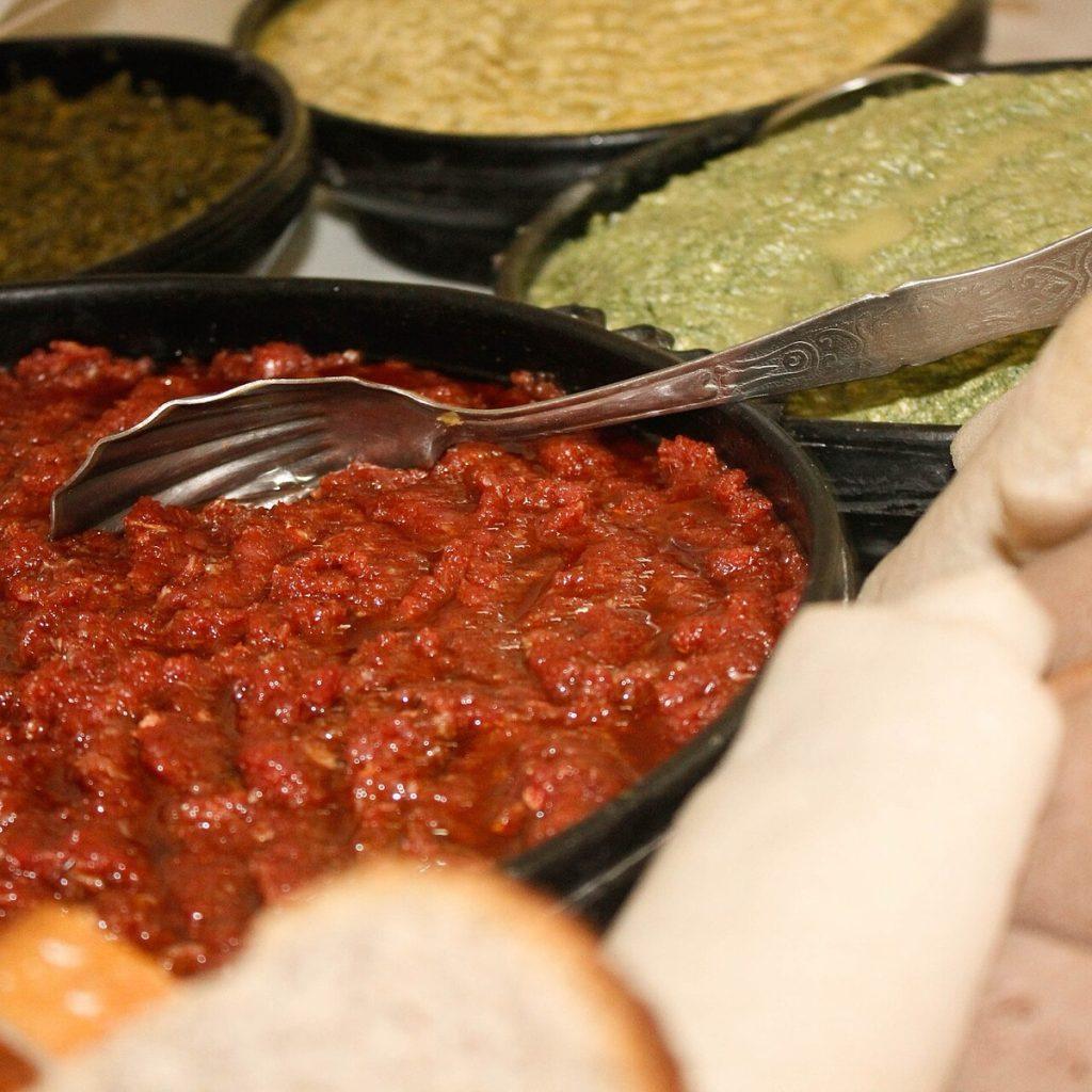 A black dish of finely minced deep red raw beef with a serving spoon, alongside dishes of greens and pale cheese and rolls of injera