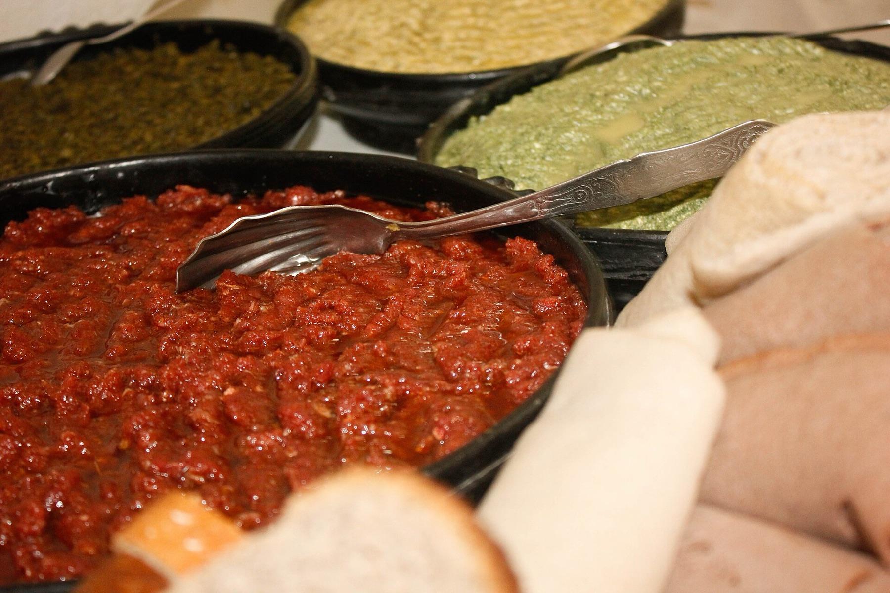 A black dish of finely minced deep red raw beef with a serving spoon, alongside dishes of greens and pale cheese and rolls of injera