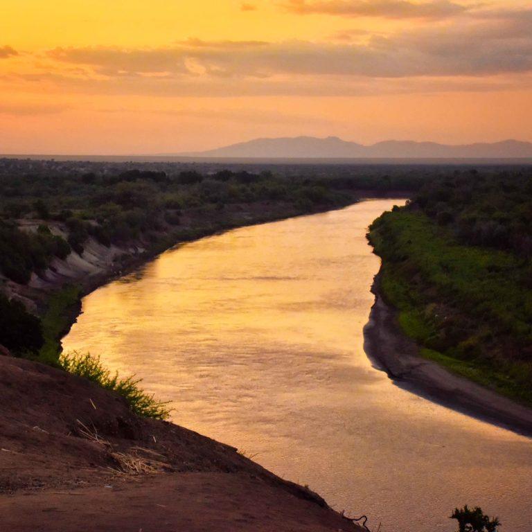 The Omo River curving through dry scrub at sunset, seen from a high bank, with low hills on the horizon