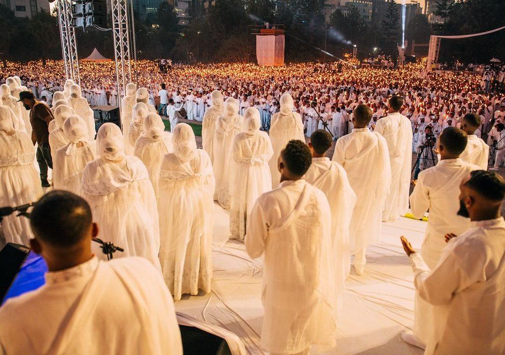 Rows of worshippers in white robes and head coverings standing on a stage in front of a huge candlelit crowd, also dressed in white, on Ethiopian Christmas Eve
