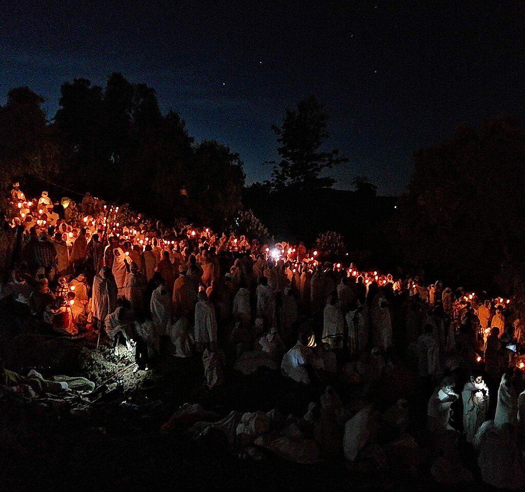 Hundreds of worshippers wrapped in white shawls holding lit candles on a hillside at night during the Genna vigil in Lalibela, with trees silhouetted against a starry sky