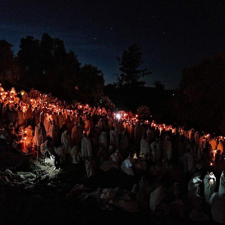 Hundreds of worshippers wrapped in white shawls holding lit candles on a hillside at night during the Genna vigil in Lalibela, with trees silhouetted against a starry sky