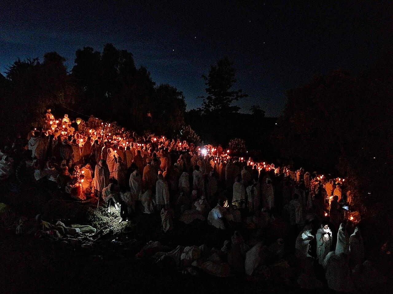 Hundreds of worshippers wrapped in white shawls holding lit candles on a hillside at night during the Genna vigil in Lalibela, with trees silhouetted against a starry sky