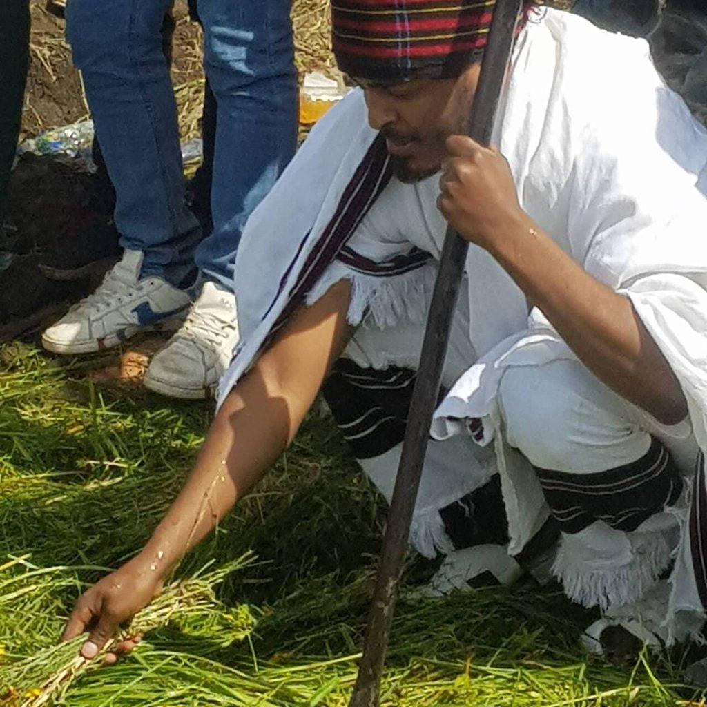 A man in a white cotton wrap and a red, black and yellow striped hat crouches holding a staff, laying a bundle of fresh green grass and small yellow flowers on the ground