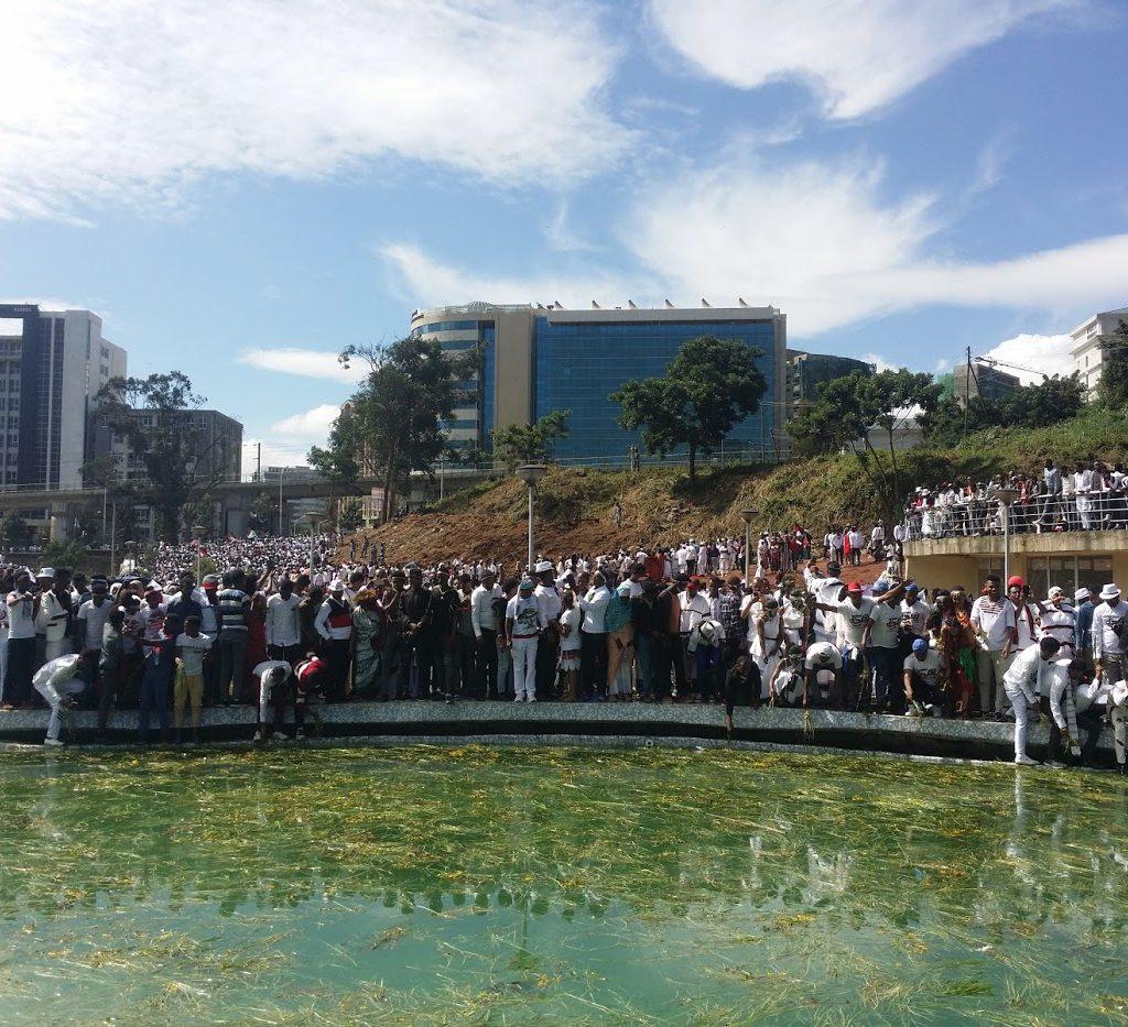 A large crowd, many in white, lines the edge of a pool in Addis Ababa whose water is covered in floating green grass, with office blocks and a hillside of people behind