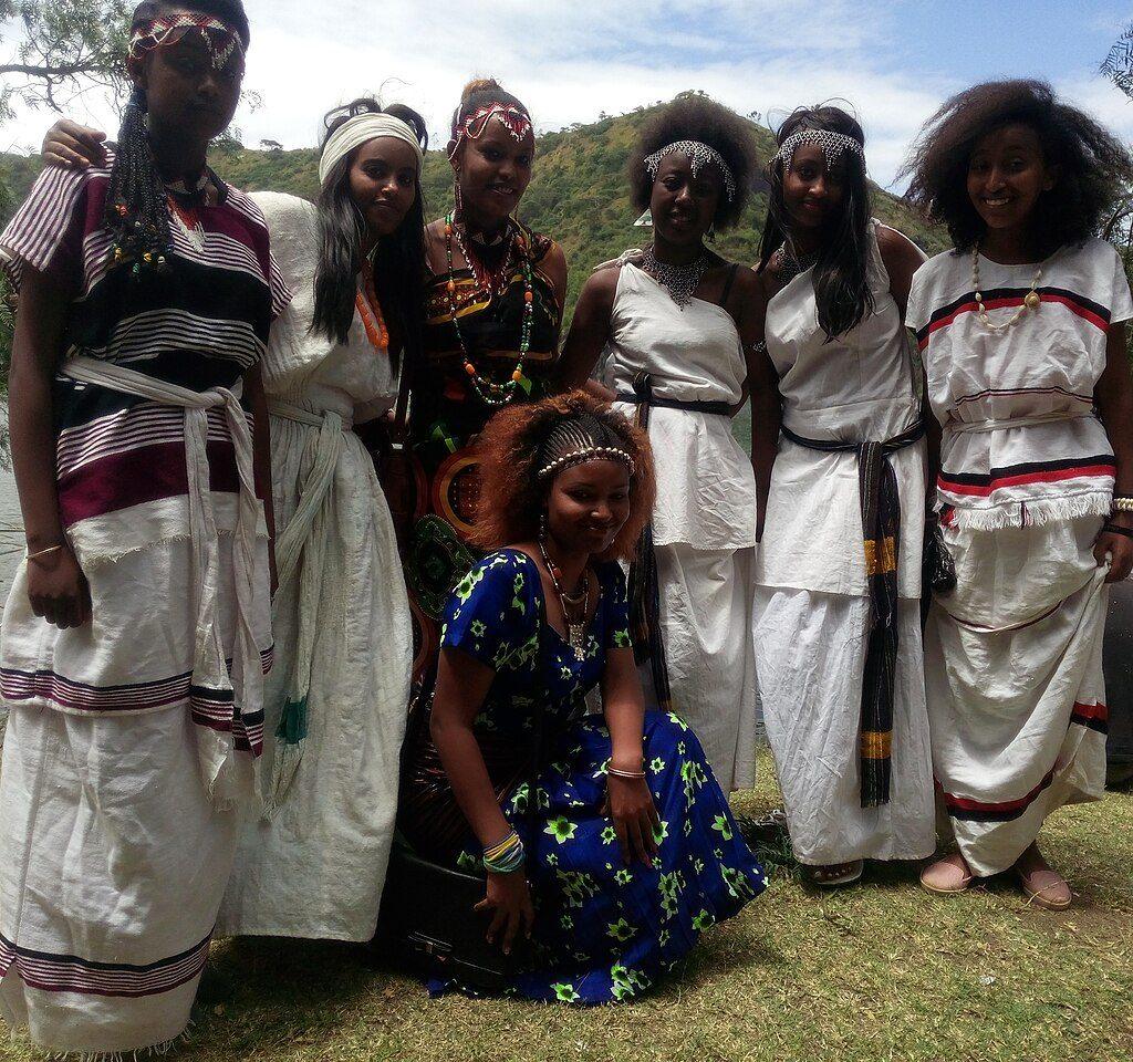 Six young women in Oromo dress, white cotton with coloured stripes and beaded headbands and necklaces, smiling together on the grassy shore of a crater lake with green hills behind