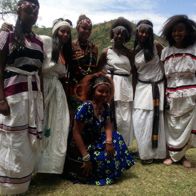 Six young women in Oromo dress, white cotton with coloured stripes and beaded headbands and necklaces, smiling together on the grassy shore of a crater lake with green hills behind