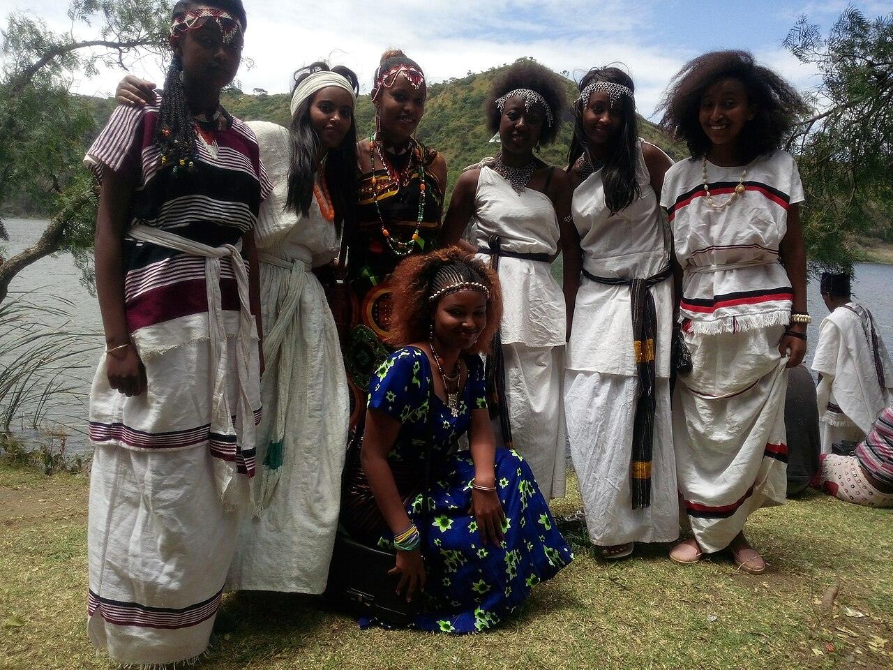 Six young women in Oromo dress, white cotton with coloured stripes and beaded headbands and necklaces, smiling together on the grassy shore of a crater lake with green hills behind
