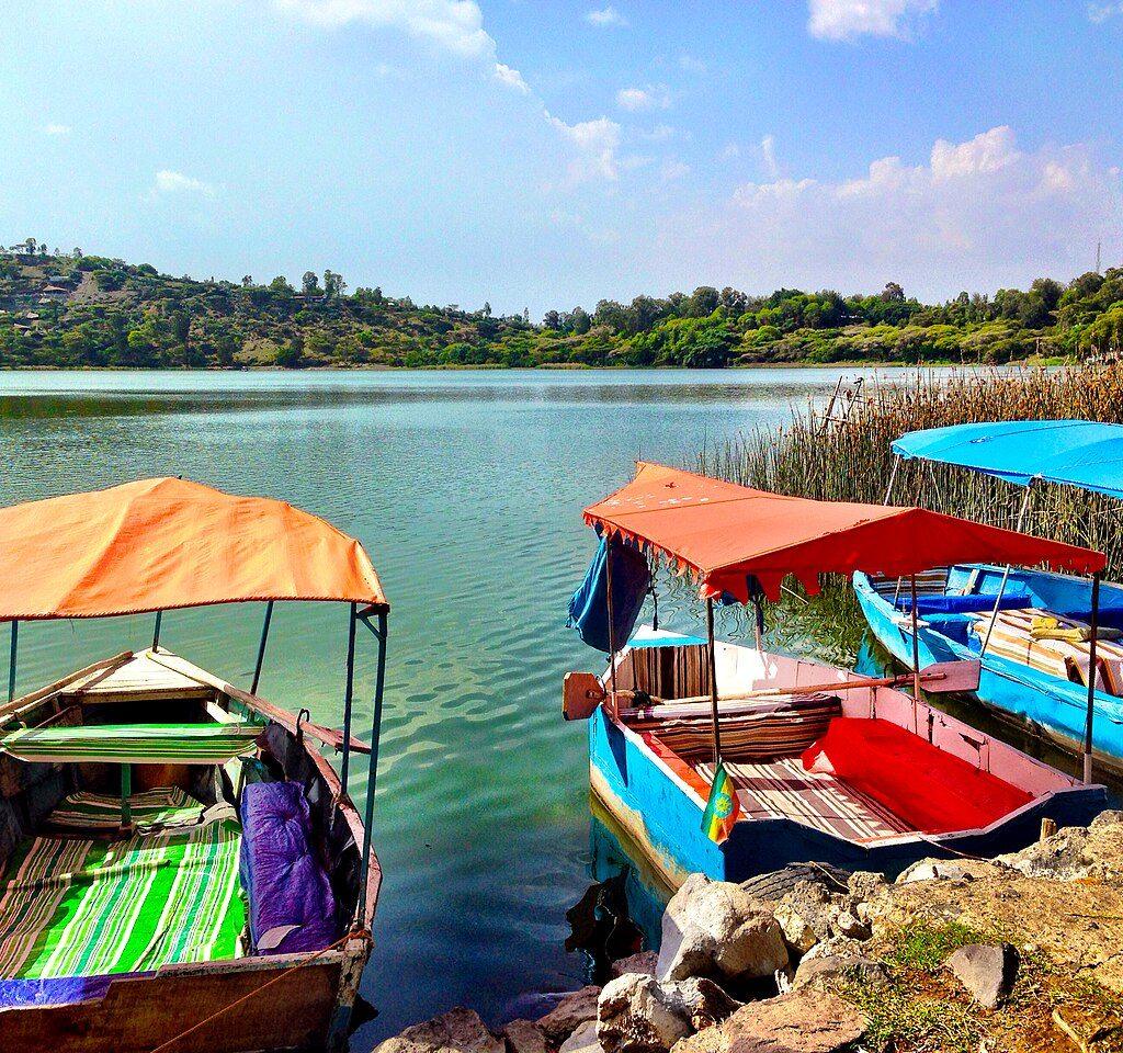 Three wooden boats with orange and blue canopies moored at the rocky shore of Lake Hora, with calm green water and wooded hills around the crater