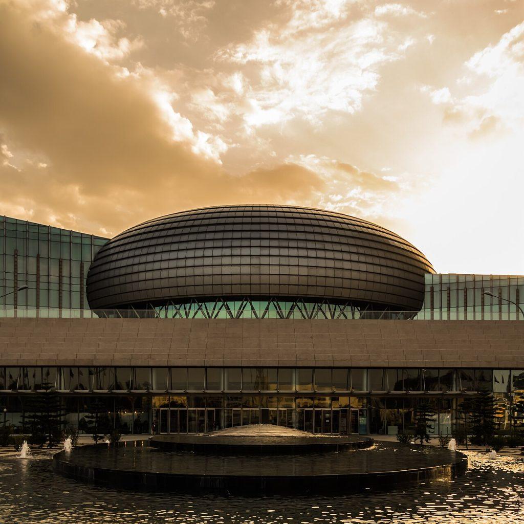 The African Union Conference Centre in Addis Ababa, a large dark oval dome rising above a long glass-fronted building, with a fountain pool in front under a golden evening sky