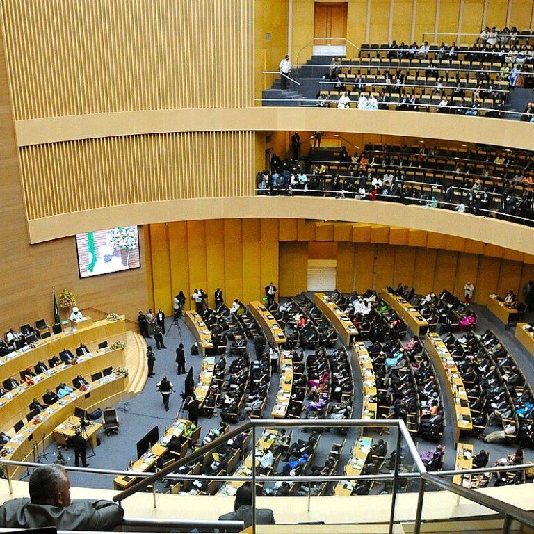 The African Union plenary chamber in Addis Ababa during a summit, with curved rows of delegates' desks below, two tiers of packed galleries and large screens on the wood-panelled walls