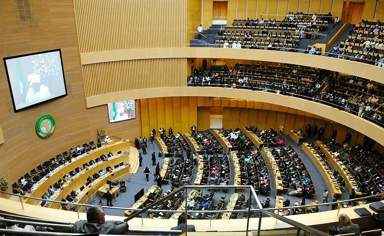 The African Union plenary chamber in Addis Ababa during a summit, with curved rows of delegates' desks below, two tiers of packed galleries and large screens on the wood-panelled walls