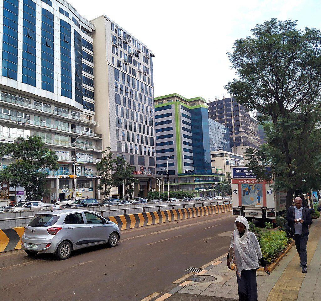 Bole Road in Addis Ababa, with tall glass-fronted hotel and office buildings, a hotel entrance, parked cars and people walking on a tree-lined pavement