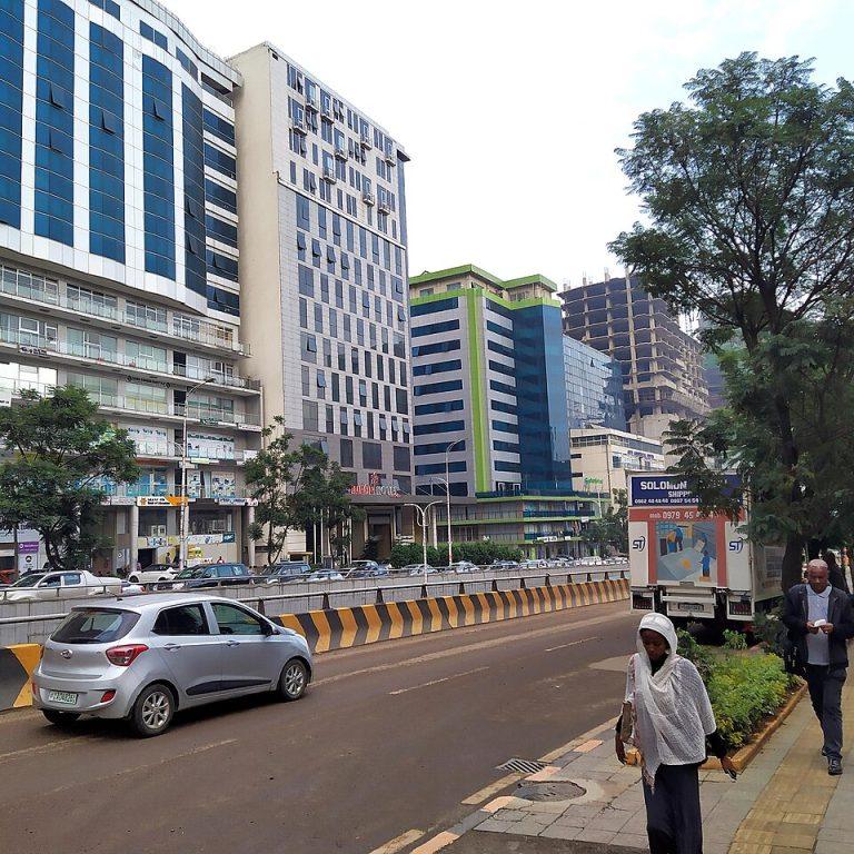 Bole Road in Addis Ababa, with tall glass-fronted hotel and office buildings, a hotel entrance, parked cars and people walking on a tree-lined pavement