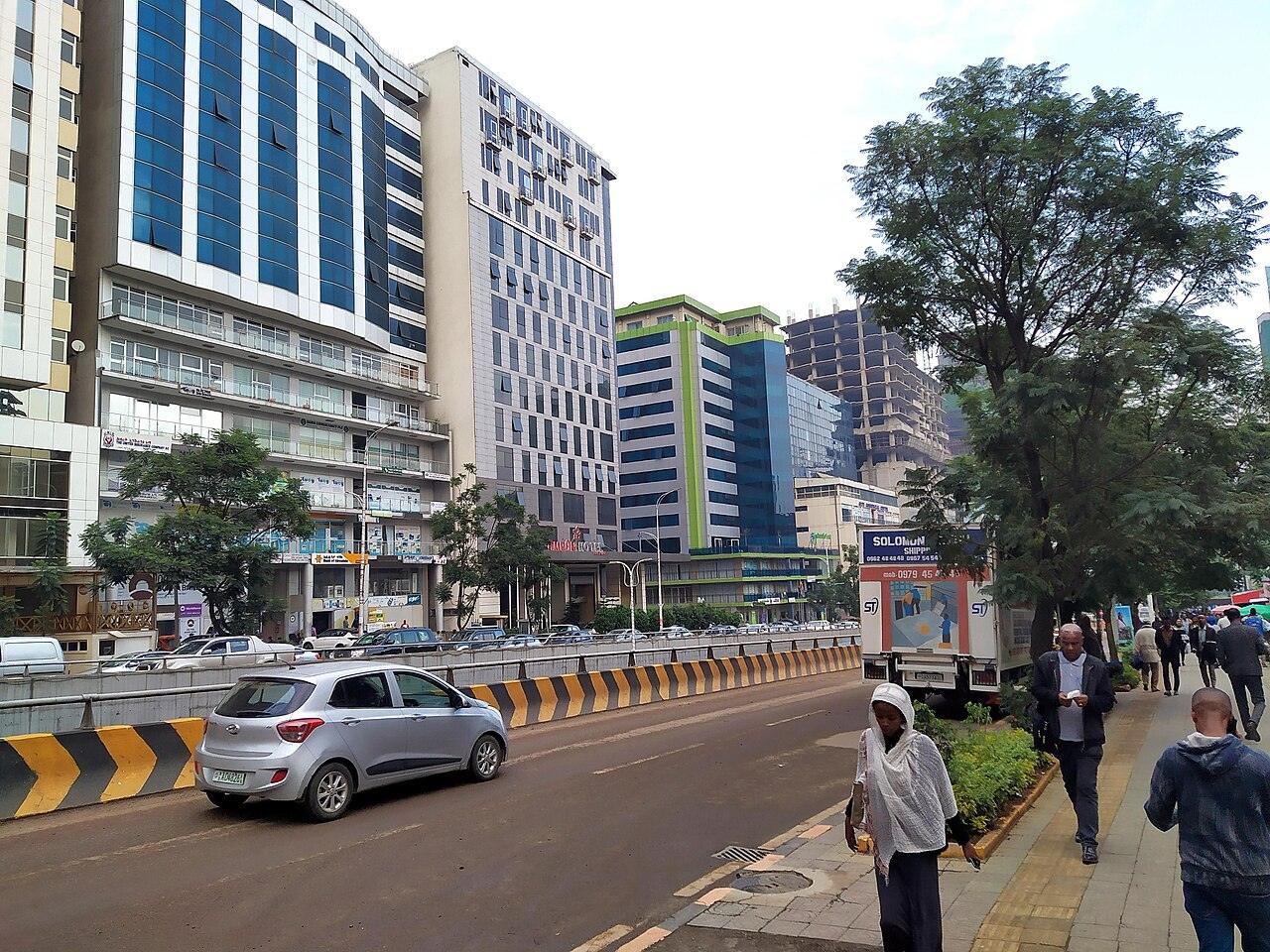 Bole Road in Addis Ababa, with tall glass-fronted hotel and office buildings, a hotel entrance, parked cars and people walking on a tree-lined pavement