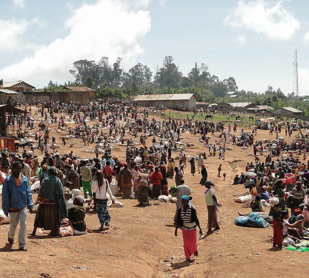 A crowded open-air market on red earth at Chencha, with hundreds of people trading among goods laid on the ground and tin-roofed buildings behind