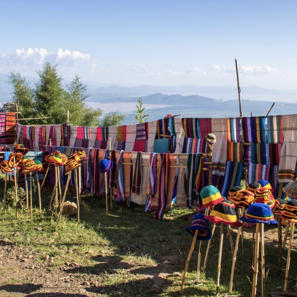 Hand-woven striped cotton shawls hung on bamboo rails and knitted hats on sticks along a hillside road, with the Rift Valley lakes far below