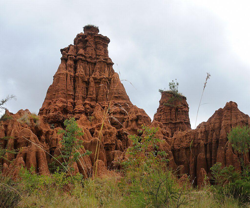 Tall eroded red sandstone pinnacles with deeply fluted sides rising above green scrub under a grey sky