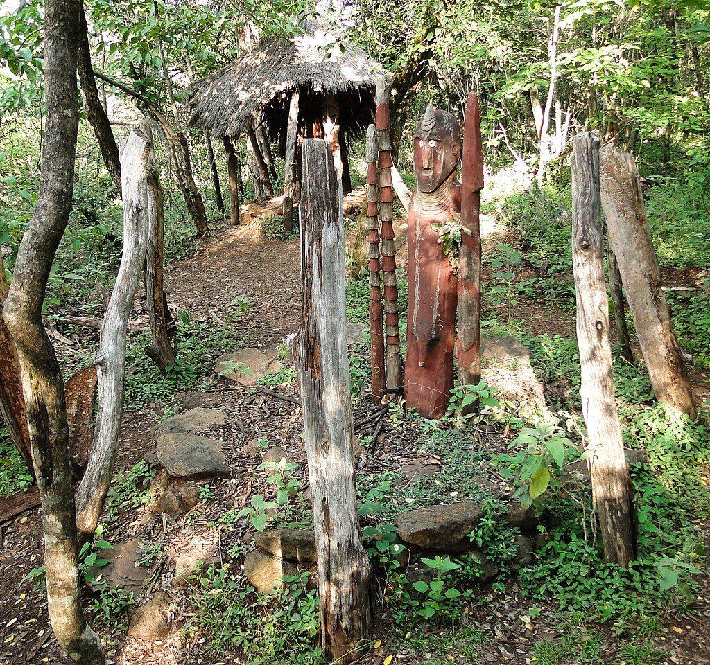 A carved wooden waga figure with a painted face and a horn-shaped headpiece standing among wooden posts in a stone-edged grave enclosure, with a thatched shelter in the woodland behind
